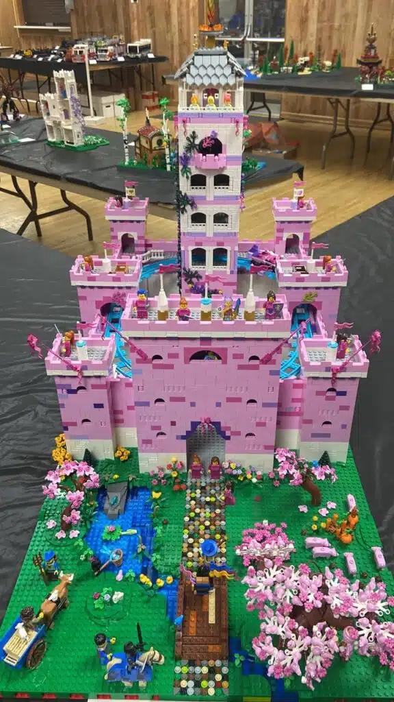 Phoebe Arcilla's "Lyserod Schloss" Version 1, showing a tall pink and white castle with many parapets and towers, and a central tall white tower.  A train on a circular blue track is built into the center of the castle.  Minifigures occupy the castle and parade on a green field out front.