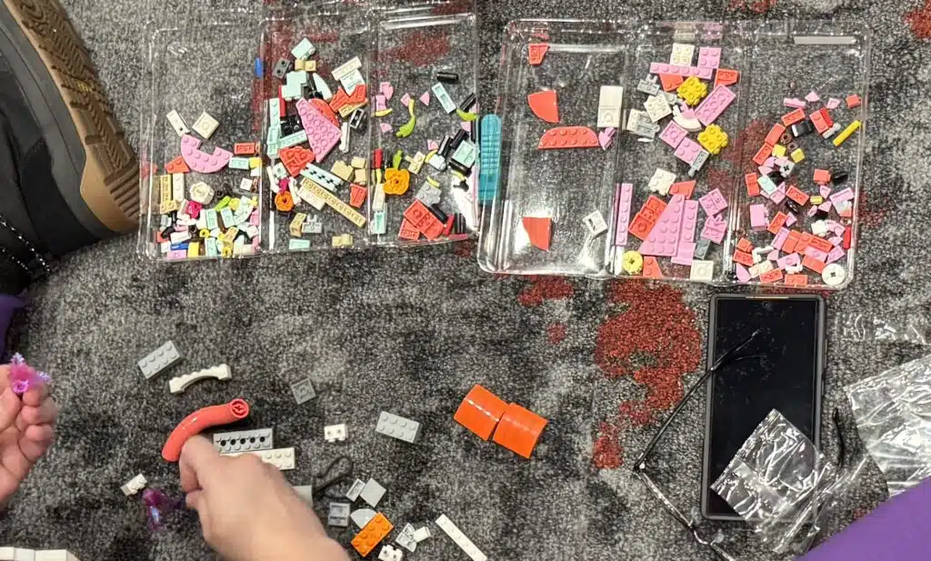 A top-down photograph shows a workshop attendee sitting on the floor, inspecting a brightly colored LEGO piece, with a variety of pink, coral, white, yellow, and light aqua parts in a plastic tray in front of them.